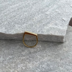 Könnte beinhalten: Ein goldfarbener Ring mit einem einzigartigen, geometrischen Design. Der Ring besteht aus einem dünnen Metallband und hat eine rechteckige Form mit einer leichten Biegung an der Oberseite.