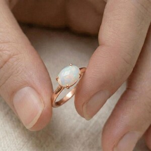 Puede incluir: Un anillo de oro rosa con una piedra preciosa de ópalo de corte ovalado. El ópalo es de color blanco lechoso con sutiles destellos iridiscentes. El anillo tiene una banda delicada de oro rosa pulido y está sujeto por cuatro garras.