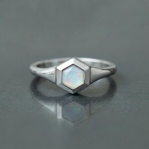 May include: A silver ring featuring a hexagonal opal gemstone. The opal has a milky white color with subtle iridescent flashes. The ring's band is smooth and polished, with a simple, elegant design.