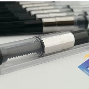 Könnte beinhalten: Nahaufnahme einer transparenten Plastik-Tintenpatrone für einen Füllfederhalter. Die Patrone ist mit schwarzer Tinte gefüllt und hat eine silberne Metallspitze. Die Patrone liegt auf einer weißen Oberfläche.