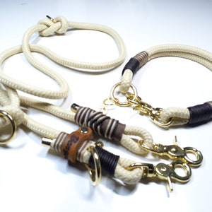 May include: A white rope dog leash and collar set with brown leather accents and gold metal hardware. The leash has a knot at the top and the collar has a brown leather tag with the word "Beast" engraved on it.