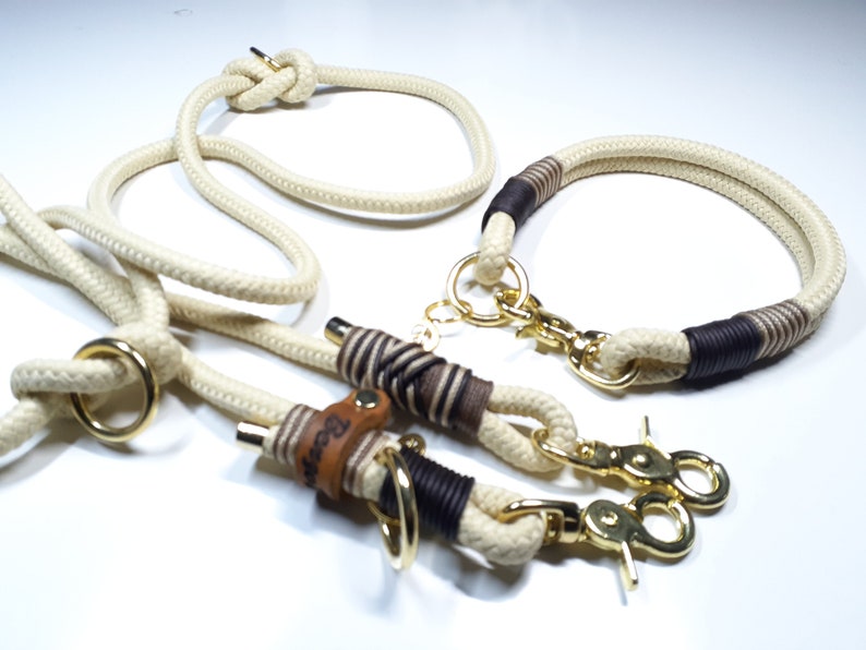 May include: A white rope dog leash and collar set with brown leather accents and gold metal hardware. The leash has a knot at the top and the collar has a brown leather tag with the word "Beast" engraved on it.