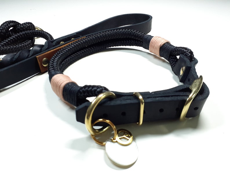 May include: Black leather dog collar with a rope leash and a gold peace sign charm. The collar has a buckle closure and the leash is made of black rope with a pink accent.