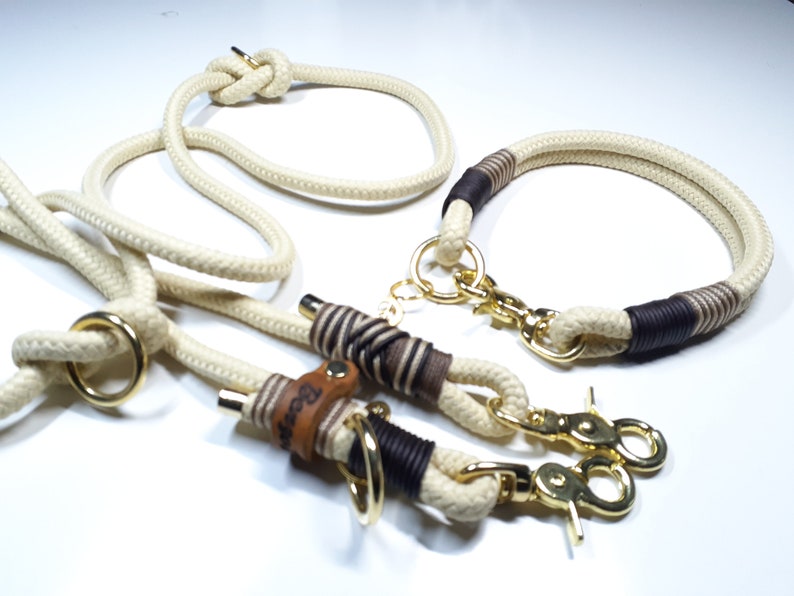 May include: A white rope dog leash and collar set with brown leather accents and gold metal hardware. The leash has a loop at one end and a clip at the other. The collar has a clip and a D-ring. The leather accents have the word "Bear" embossed on them.