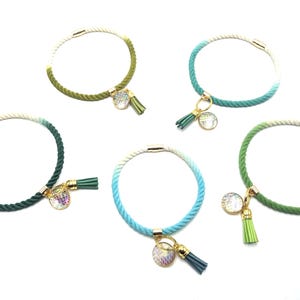 Könnte beinhalten: Fünf farbige Seilarmbänder mit goldfarbenen Verschlüssen und Anhängern. Jedes Armband hat einen Farbverlauf, mit einer Quaste und einem runden, irisierenden Anhänger. Die Farben umfassen Grün und Blau.
