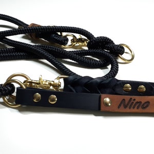 May include: Black leather dog collar with a brown leather name tag that says "Nino" and gold metal rivets. The collar is attached to a black rope leash with a gold metal clip.