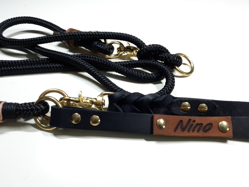 May include: Black leather dog collar with a brown leather name tag that says "Nino" and gold metal rivets. The collar is attached to a black rope leash with a gold metal clip.