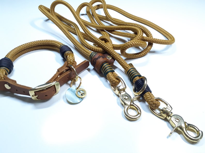 Puede incluir: Un collar de perro de cuero marr&oacute;n con una hebilla dorada y un dije de signo de paz, combinado con una correa de cuerda marr&oacute;n y verde a juego con dos cierres dorados.