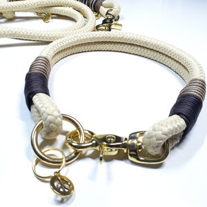 May include: A white rope dog collar with brown and black accents. The collar has a gold metal clasp and a peace sign charm.