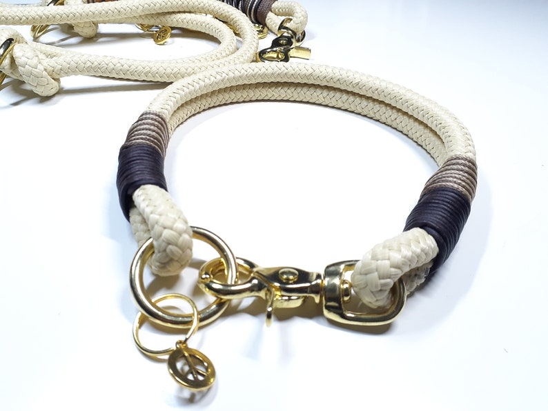 May include: A white rope dog collar with brown and black accents. The collar has a gold metal clasp and a peace sign charm.
