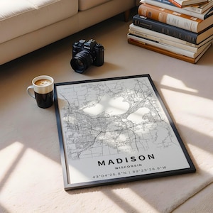 May include: A framed map print of Madison, Wisconsin, featuring black street lines on a white background. The text reads MADISON WISCONSIN 43&deg;04'25.8"N | 89&deg;23'28.9"W. The print is displayed alongside a black camera and a white ceramic coffee mug.