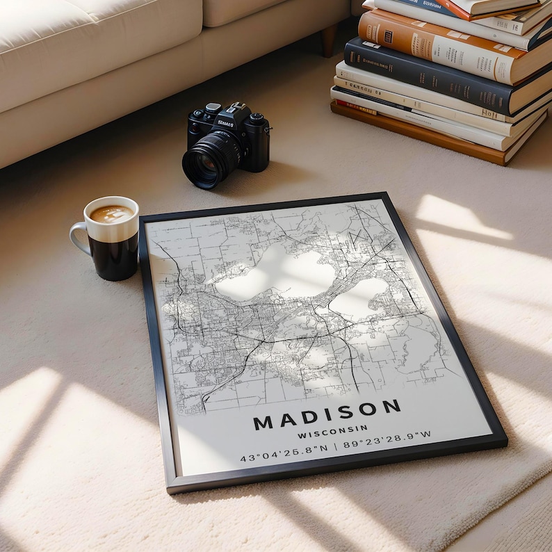 May include: A framed map print of Madison, Wisconsin, featuring black street lines on a white background. The text reads MADISON WISCONSIN 43&deg;04'25.8"N | 89&deg;23'28.9"W. The print is displayed alongside a black camera and a white ceramic coffee mug.