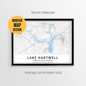 May include: A framed map print of Lake Hartwell in Georgia and South Carolina featuring light blue water, thin black road lines, and the text "LAKE HARTWELL GEORGIA & SOUTH CAROLINA 34°27'34.5"N | 82°51'38.7"W" printed in black at the bottom of the image.