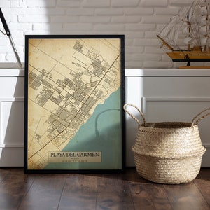 Puede incluir: Una impresi&oacute;n de mapa de estilo vintage de Playa del Carmen, M&eacute;xico, en un marco negro. El mapa est&aacute; impreso en papel envejecido con un esquema de color azul y verde. El mapa se muestra en un suelo de madera con una cesta tejida y un velero modelo en el fondo.