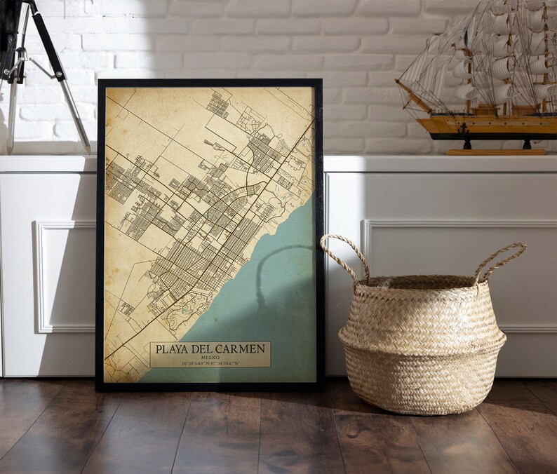 Puede incluir: Una impresi&oacute;n de mapa de estilo vintage de Playa del Carmen, M&eacute;xico, en un marco negro. El mapa est&aacute; impreso en papel envejecido con un esquema de color azul y verde. El mapa se muestra en un suelo de madera con una cesta tejida y un velero modelo en el fondo.