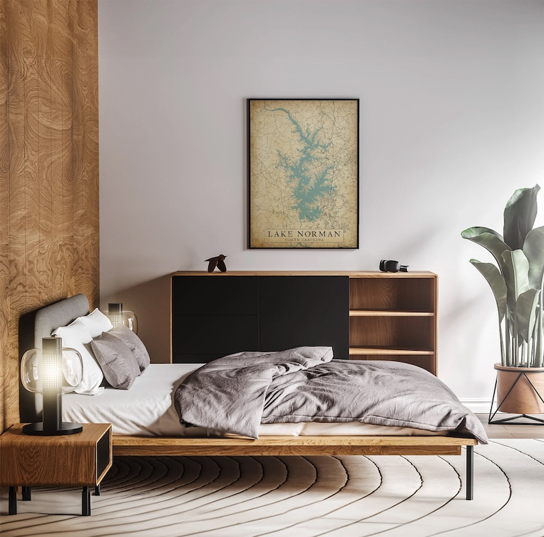 Puede incluir: Un dormitorio moderno con una cama de madera, una colcha gris y una mesita de noche con una l&aacute;mpara. Un mapa enmarcado del lago Norman, Carolina del Norte, cuelga de la pared. La habitaci&oacute;n tiene un dise&ntilde;o moderno con colores neutros y una gran planta en maceta.