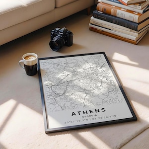 May include: A framed black and white map of Athens, Georgia, with the city name and coordinates printed at the bottom. A black camera, a coffee mug, and a stack of books are also visible.