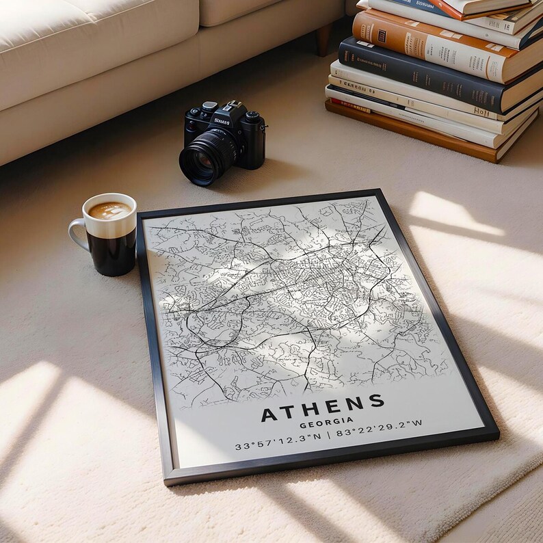 May include: A framed black and white map of Athens, Georgia, with the city name and coordinates printed at the bottom. A black camera, a coffee mug, and a stack of books are also visible.