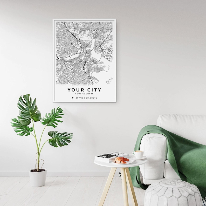 May include: A framed black and white map print with the text "YOUR CITY" and "YOUR COUNTRY" at the bottom. The map shows a detailed road network. A potted plant with large green leaves is in the foreground.
