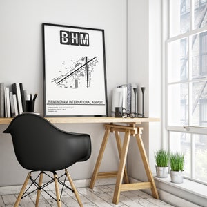 May include: Black and white print of Birmingham International Airport (BHM) layout with the airport code "BHM" in a black box at the top. The print is framed and hanging on a white wall above a desk with a black chair and a wooden easel.