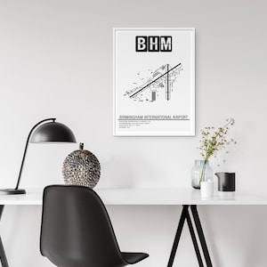 May include: A black and white print of Birmingham International Airport's layout with the airport code "BHM" at the top. The print is framed in a white frame and is hanging on a white wall above a desk with a lamp, a decorative sphere, and a vase of flowers.