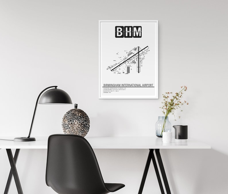 May include: A black and white print of Birmingham International Airport's layout with the airport code "BHM" at the top. The print is framed in a white frame and is hanging on a white wall above a desk with a lamp, a decorative sphere, and a vase of flowers.