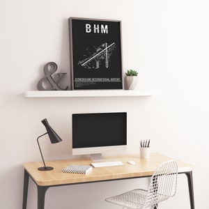 May include: Black and white print of Birmingham International Airport layout with the airport code "BHM" at the top. The print is framed and hanging on a white shelf above a desk with a computer, lamp, and chair.