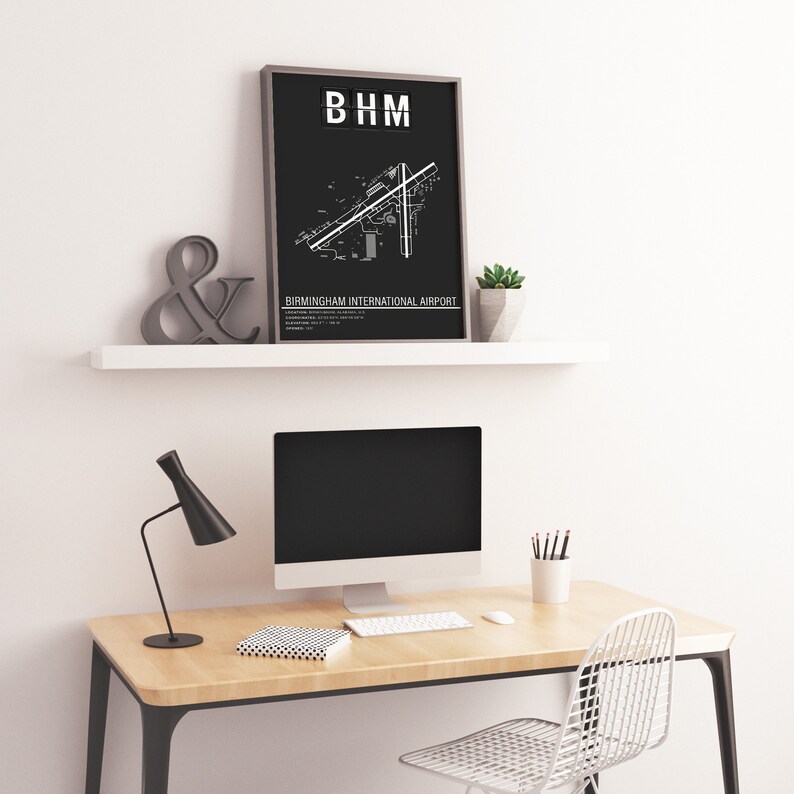 May include: Black and white print of Birmingham International Airport layout with the airport code "BHM" at the top. The print is framed and hanging on a white shelf above a desk with a computer, lamp, and chair.