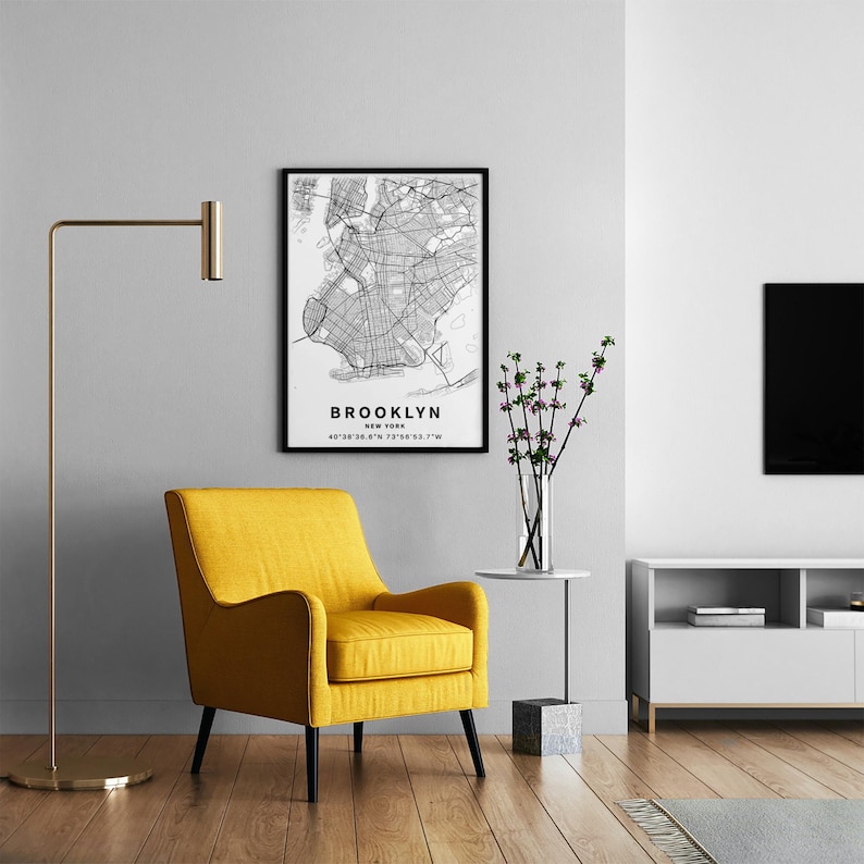May include: A mustard yellow upholstered armchair with black legs, positioned next to a tall gold floor lamp and a small side table holding a glass vase with green branches. A framed black and white map print of Brooklyn, New York hangs above.