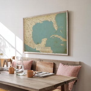Puede incluir: Un mapa enmarcado del Golfo de Am&eacute;rica, con un mar azul claro y masas terrestres beige. El mapa est&aacute; en un marco blanco y cuelga sobre un banco de madera con cojines a cuadros rojos y blancos. El texto "GULF OF AMERICA" es visible.
