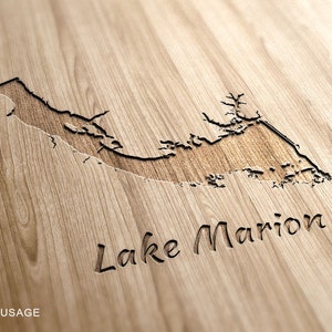 May include: Wooden plaque with the engraved outline of Lake Marion. The lake's shape is carved into the wood, with the name "Lake Marion" below. The wood has a natural grain and a warm, light brown color.