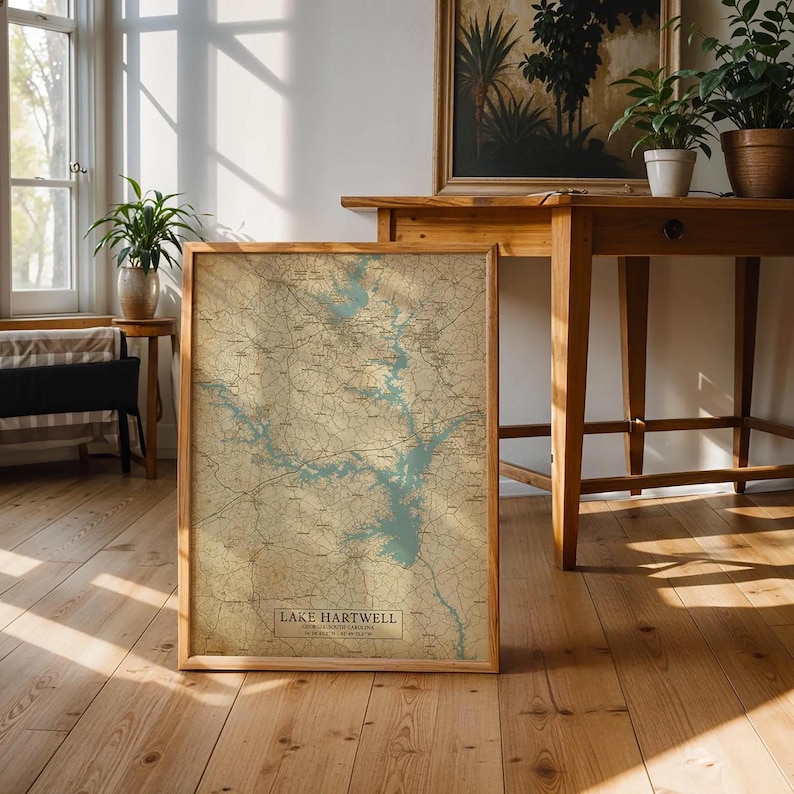 Puede incluir: Un mapa enmarcado del lago Hartwell, con un dise&ntilde;o topogr&aacute;fico detallado en tonos beige y azul. El mapa est&aacute; en un marco de madera y descansa sobre un suelo de madera. El texto "Lake Hartwell" es visible en la parte inferior.