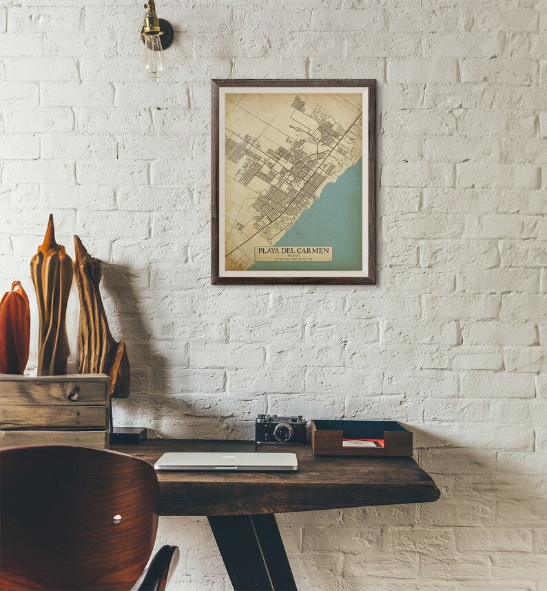 Puede incluir: Un mapa vintage enmarcado de Playa del Carmen, M&eacute;xico, con un esquema de color azul y beige. El mapa est&aacute; colgado en una pared de ladrillo blanco sobre un escritorio de madera con una computadora port&aacute;til, una c&aacute;mara y un organizador de madera.