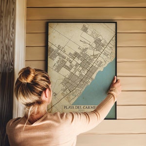 Puede incluir: Una mujer cuelga un mapa de estilo vintage de Playa del Carmen, M&eacute;xico, en una pared de madera. El mapa est&aacute; impreso en papel envejecido con l&iacute;neas y texto negros. El mapa est&aacute; enmarcado en un marco negro.