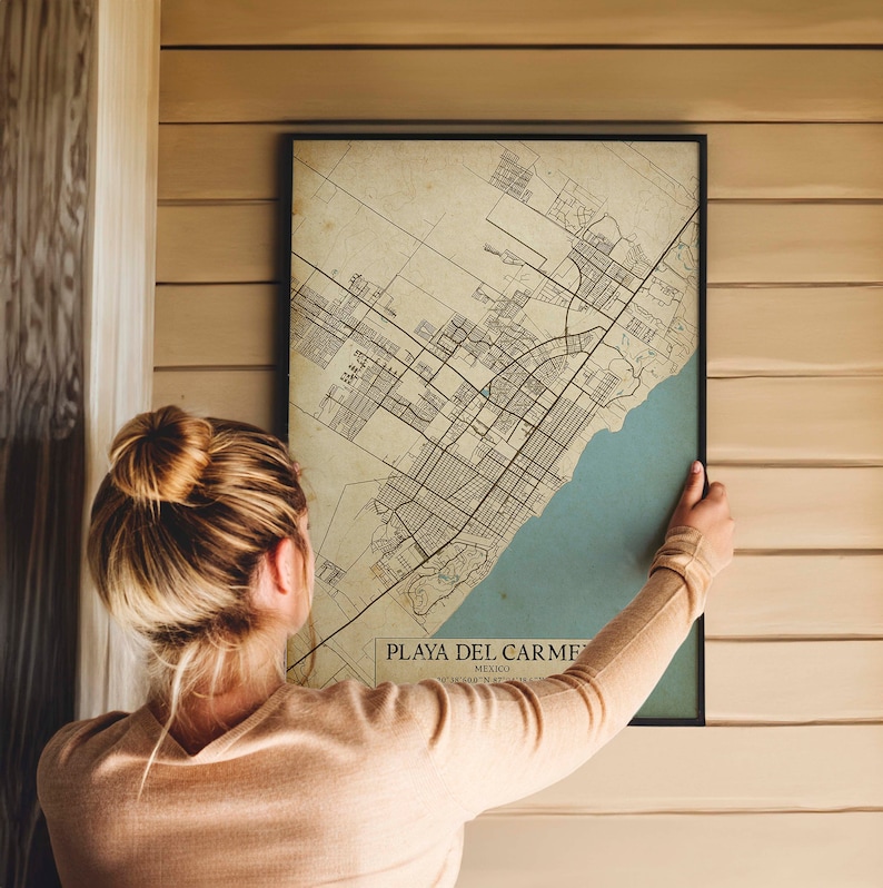 Puede incluir: Una mujer cuelga un mapa de estilo vintage de Playa del Carmen, M&eacute;xico, en una pared de madera. El mapa est&aacute; impreso en papel envejecido con l&iacute;neas y texto negros. El mapa est&aacute; enmarcado en un marco negro.