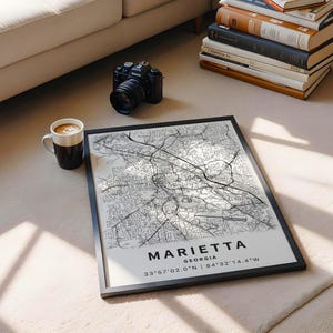 Może przedstawiać: Oprawiona mapa Marietta w stanie Georgia z czarnymi liniami ulic na białym tle. Tekst głosi MARIETTA GEORGIA 33&deg;57'02.0"N | 84&deg;32'14.4"W. Wydruk jest pokazany obok czarnego aparatu cyfrowego oraz białego kubka do kawy.