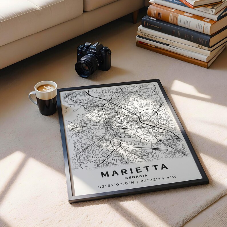 Może przedstawiać: Oprawiona mapa Marietta w stanie Georgia z czarnymi liniami ulic na białym tle. Tekst głosi MARIETTA GEORGIA 33&deg;57'02.0"N | 84&deg;32'14.4"W. Wydruk jest pokazany obok czarnego aparatu cyfrowego oraz białego kubka do kawy.