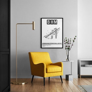 May include: A black and white airport map print with the text "BHM" and "Birmingham International Airport" in a black frame hanging on a white wall. A yellow armchair, a floor lamp, a vase with flowers, and a white TV stand are in the background.