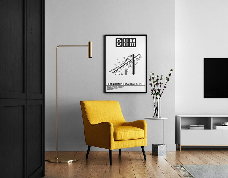 May include: A black and white airport map print with the text "BHM" and "Birmingham International Airport" in a black frame hanging on a white wall. A yellow armchair, a floor lamp, a vase with flowers, and a white TV stand are in the background.