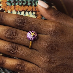 Può includere: Un anello d'oro con un designo a forma di stella marina viola e gialla. L'anello è indossato al dito di una persona.