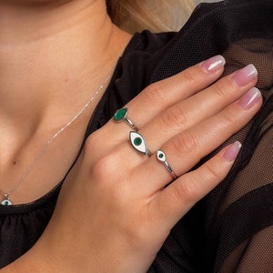 May include: A woman wearing a black top with a silver necklace and three silver rings with green evil eye designs.