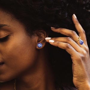 May include: A gold-toned ring featuring a small blue and white seashell accent and a matching blue and white seashell stud earring. The jewellery set displays a distinct ribbed texture on the shells, with the ring band having a wavy, organic shape.