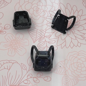 May include: Three small, black plastic backpack components are arranged on a pink floral patterned surface. The components include a backpack base, a top piece, and a backpack with straps. The items are likely for a doll or toy.