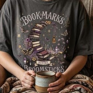 May include: A charcoal gray t-shirt featuring a graphic of a crescent moon made of stacked books, surrounded by flowers, butterflies, and crystals. The text reads "BOOKMARKS BEFORE BROOMSTICKS" in a decorative font across the chest area.
