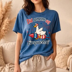 May include: A blue short-sleeve t-shirt featuring a graphic of a white duck wearing sunglasses and riding a red, white, and blue firecracker. The text reads, Did someone say firequacker? The shirt is paired with light beige, loose-fitting trousers.