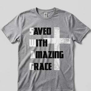 Puede incluir: Camiseta gris jaspeado con la frase "SAVED WITH AMAZING GRACE" en un diseño en capas. Un gráfico de cruz blanca está integrado en el texto. La camiseta tiene cuello redondo y mangas cortas.