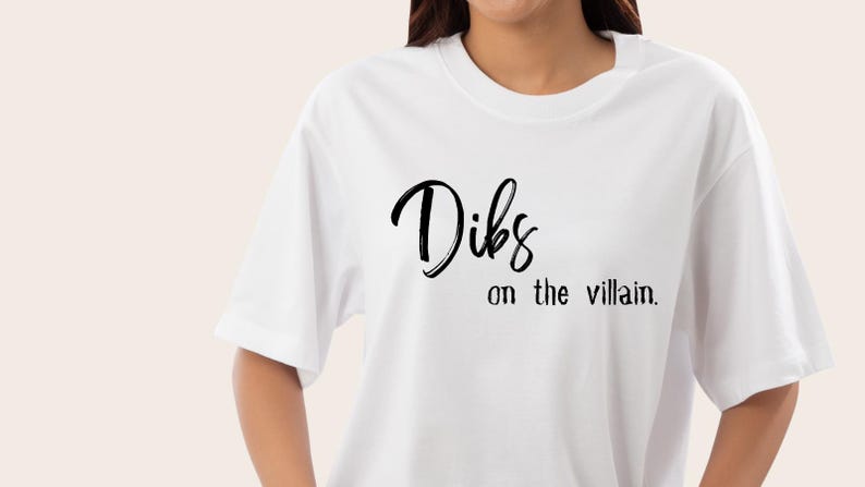May include: White t-shirt with the phrase "Dibs on the villain" in black script. The shirt has short sleeves and a crew neck. The text is centered on the front of the shirt.