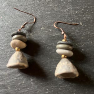 Peut inclure: Une paire de boucles d'oreilles pendantes composées de pierres empilées grises et beiges suspendues à des crochets en métal cuivré. Chaque boucle comprend deux petites pierres circulaires plates, une perle dorée et une pierre en forme de cloche.