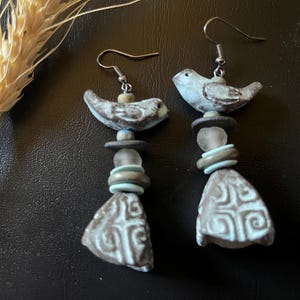 Peut inclure: Une paire de boucles d'oreilles pendantes avec un oiseau en céramique bleu clair et marron, des perles empilées bleu clair et grises, et un pendentif triangulaire en céramique orné d'un motif géométrique blanc en relief.