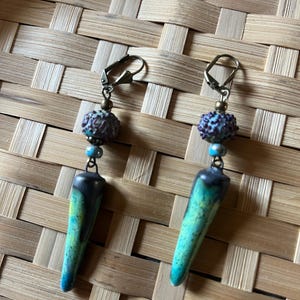 May include: A pair of dangle earrings featuring bronze-toned leverback hooks, a small round metallic bead, a textured purple spherical bead, a tiny light blue bead, and a long, tapered ceramic pendant with a green, yellow, and black glaze.